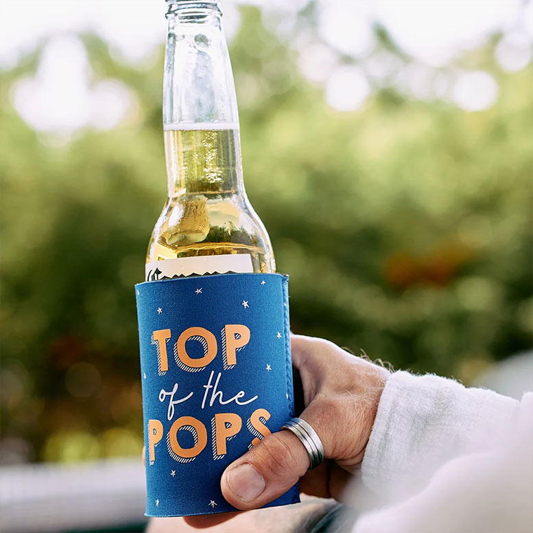 Hand holding a bottle with a 'Top of the Pops' can cooler against a blurred green background