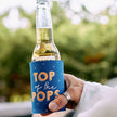 Hand holding a bottle with a 'Top of the Pops' can cooler against a blurred green background
