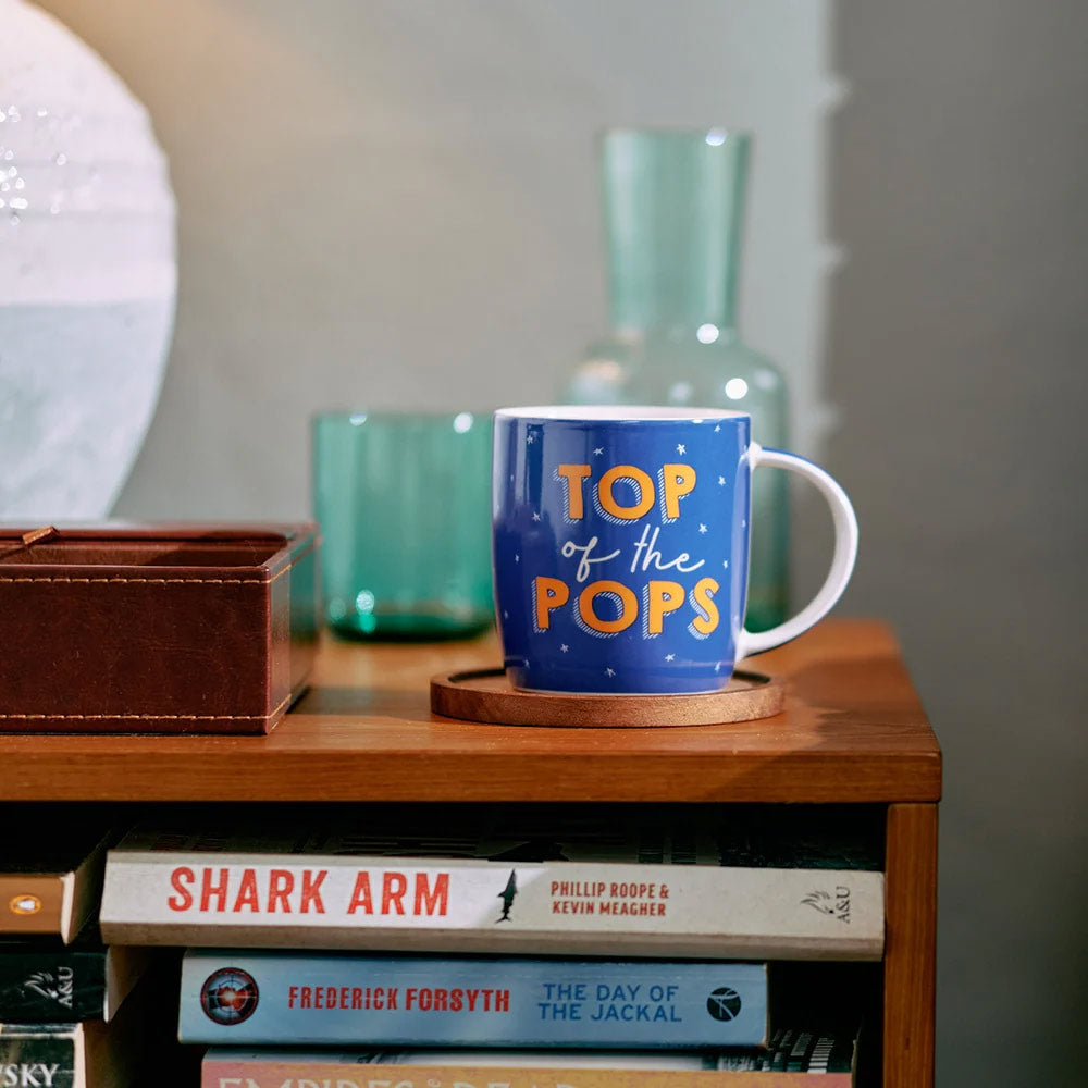 Blue mug with 'Top of the Pops' text on a wooden surface with books and a vase in the background