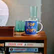 Blue mug with 'Top of the Pops' text on a wooden surface with books and a vase in the background