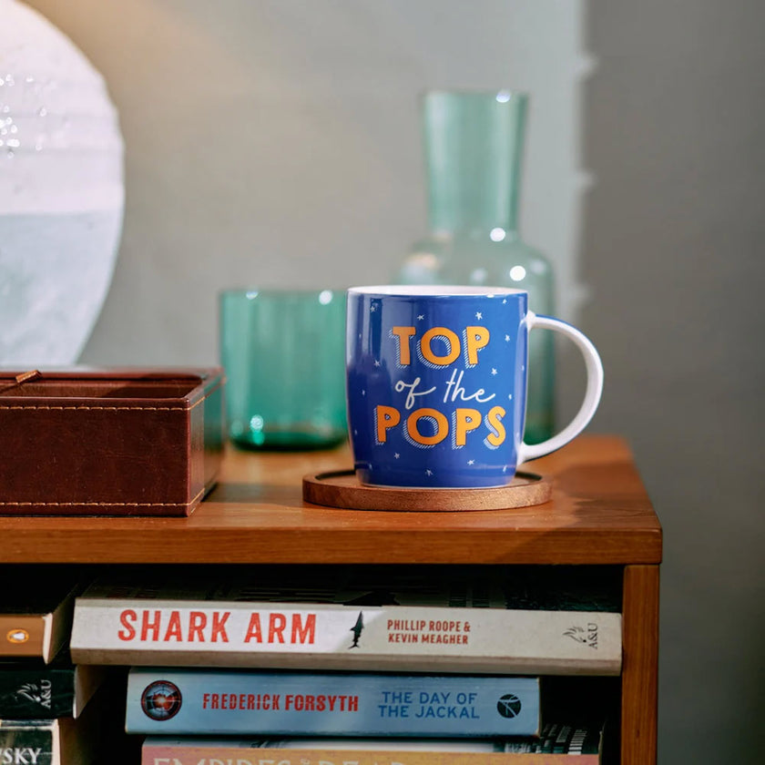 Blue mug with 'Top of the Pops' text on a wooden surface with books and a vase in the background