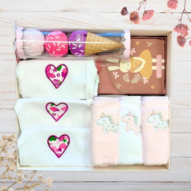 Set of folded baby clothes with heart, baby girl socks and unicorn washers in a box on a wooden surface.