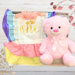 Pink teddy bear with a 'one' birthday romper in a decorative gift box on a wooden surface.