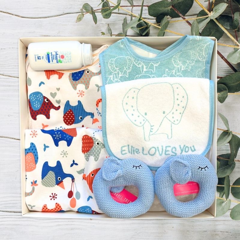Baby gift set with bibs, elephant toys, and baby powder in a gift box on a wooden surface with greenery.