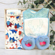 Baby gift set with bibs, elephant toys, and baby powder in a gift box on a wooden surface with greenery.