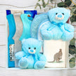 Gift set with blue teddy bears, baby clothes, and Lindt chocolate bars on a wooden surface.