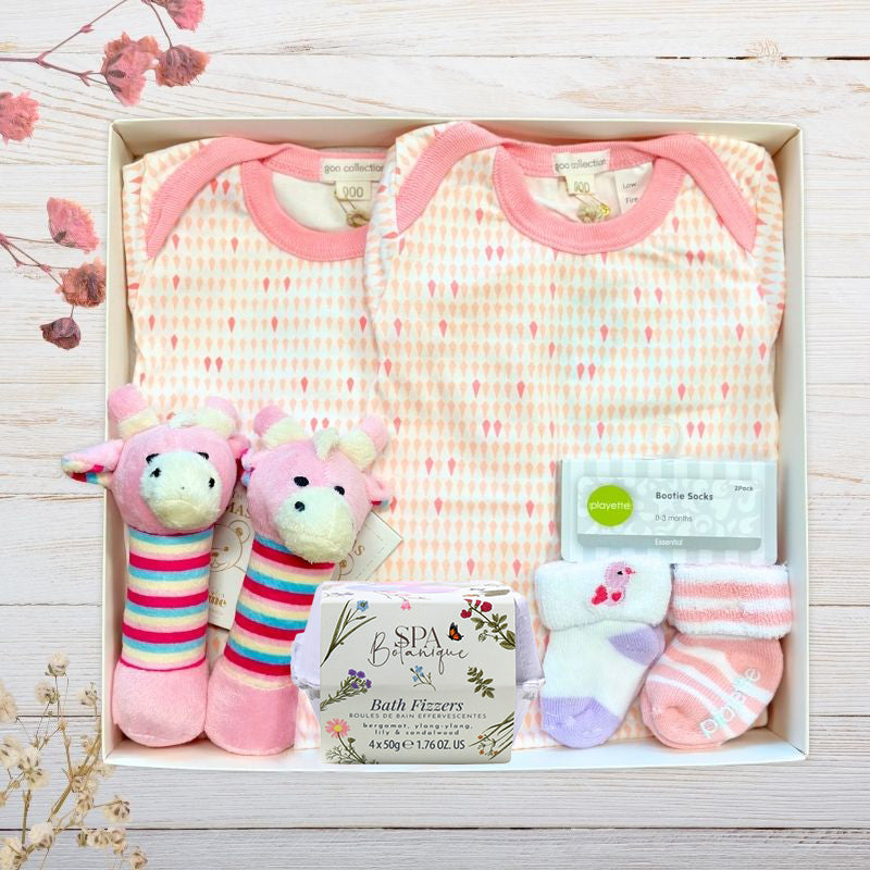 Baby gift set with twin organic pink onesies, socks, and toys in a gift box on a wooden surface.