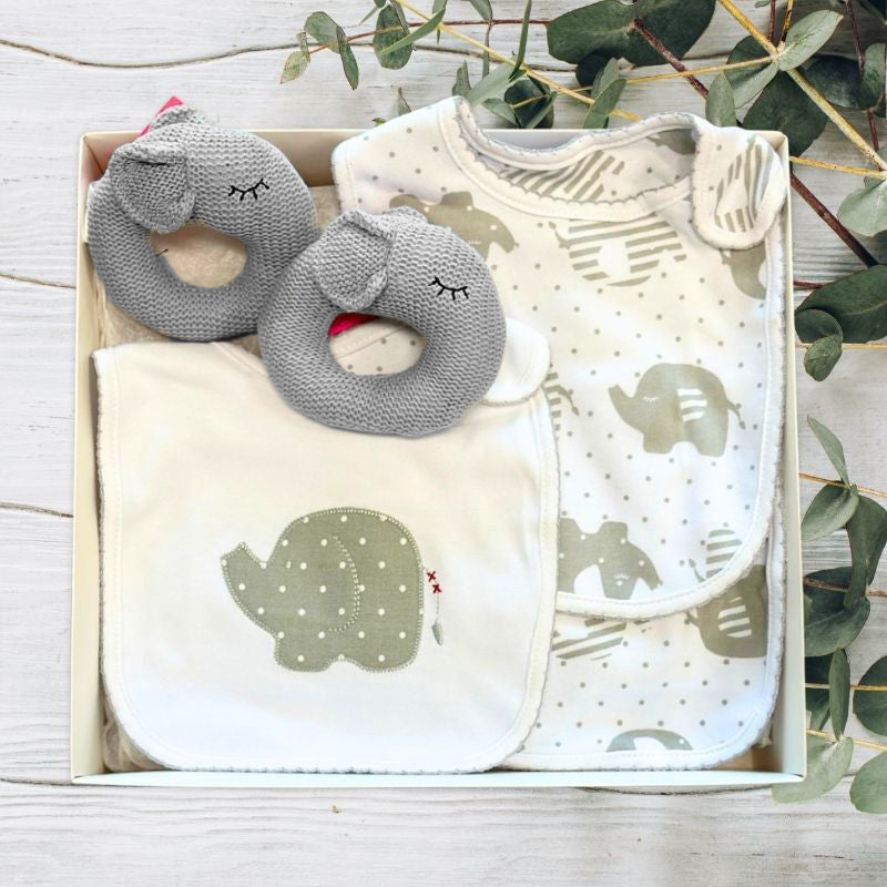 Set of baby bibs and toys with elephant patterns in a gift box on a wooden surface.