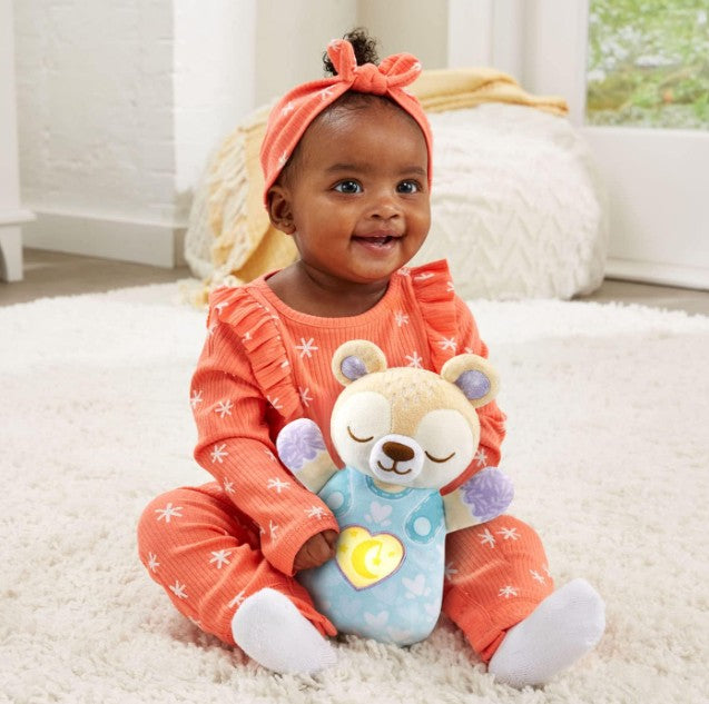 Baby in an orange outfit holding a teddy bear with a heart light on a carpeted floor.