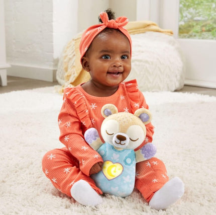 Baby in an orange outfit holding a teddy bear with a heart light on a carpeted floor.