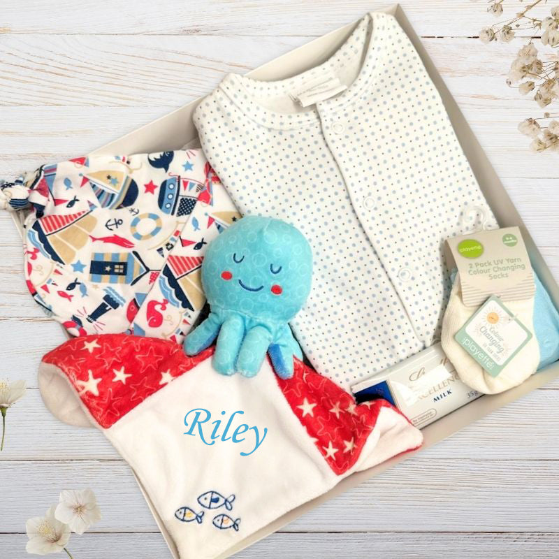 Baby gift set with blue octopus blanket, baby clothes, and other items in a gift box on a light wooden surface.
