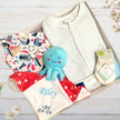 Baby gift set with blue octopus blanket, baby clothes, and other items in a gift box on a light wooden surface.