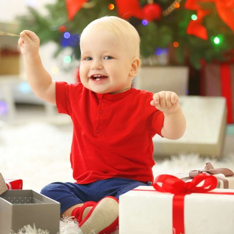 Child in a red shirt sitting among Christmas presents with a decorated tree in the background