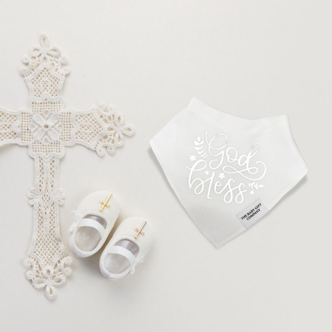 White cross, baby shoes, and bib with 'God bless' text on a light gray background