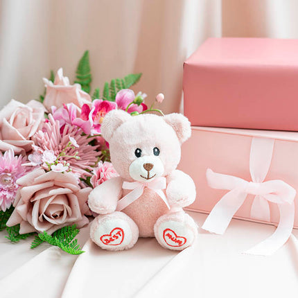 White teddy bear with pink accents, flowers, and a gift box on a light surface.