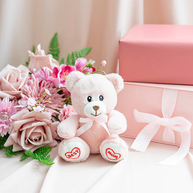 White teddy bear with pink accents, flowers, and a gift box on a light surface.