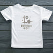 White t-shirt with '1st Birthday Boy' text on a wooden surface
