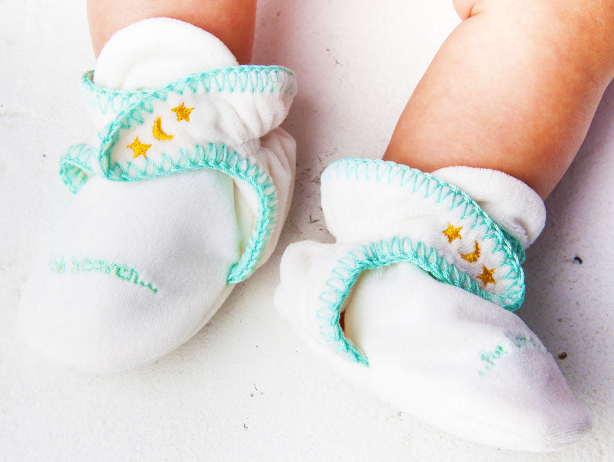 Baby feet wearing white slippers with teal ribbing and star patterns on a light background