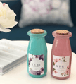 Two candle bottles with floral labels and cork lids on a surface with a blurred background.