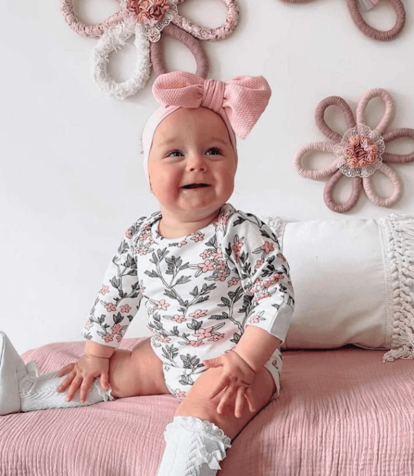Baby wearing a floral onesie and pink headband sitting on a pink surface with decorative flowers in the background.