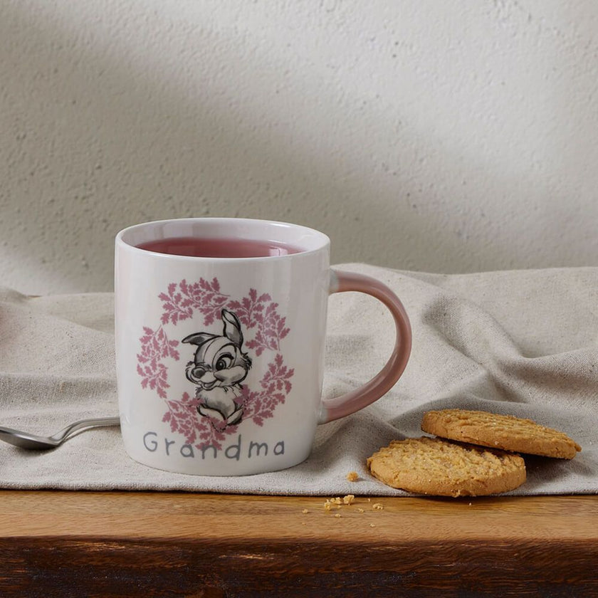 White mug with pink handle featuring a design and the word 'Grandma', filled with a red beverage, on a wooden surface with cookies and a spoon.