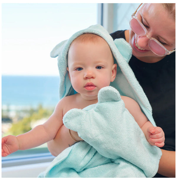 Baby wrapped in a light green towel with ears, held by an adult outdoors.