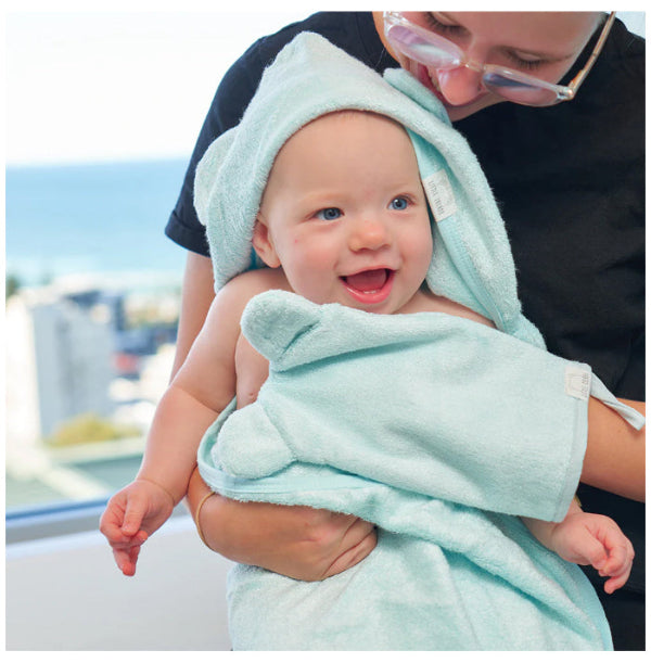 Baby wrapped in a light green towel with a hood, held by an adult outdoors.