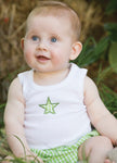 Baby wearing a white sleeveless shirt with a green star and green checkered shorts sitting outdoors.