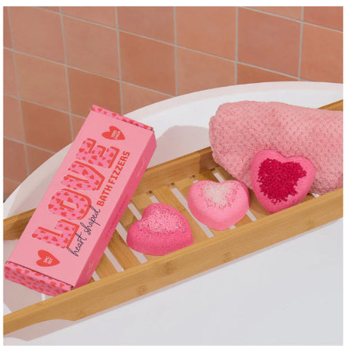 Pink bath fizzer packaging with heart-shaped bath bombs on a wooden tray against a tiled bathroom wall.