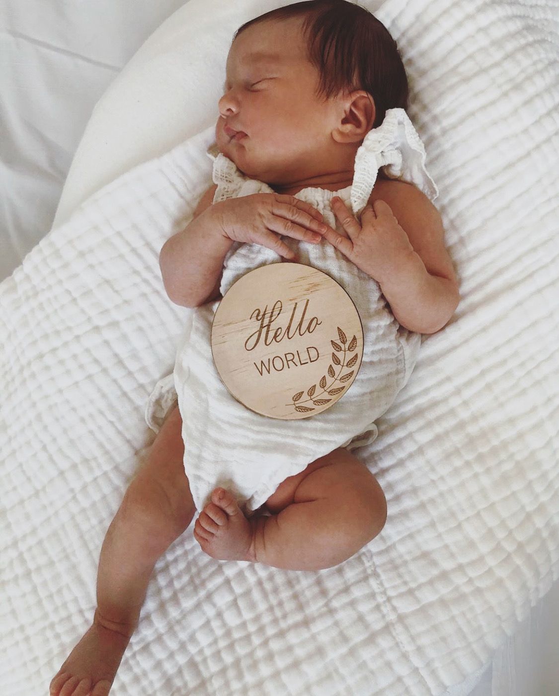 Newborn baby lying on a white blanket with a wooden disc that says 'Hi Hello World'.