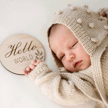Newborn baby in a beige knitted outfit with a 'Hello World' sign on a white background
