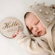 Newborn baby in a beige knitted outfit with a 'Hello World' sign on a white background