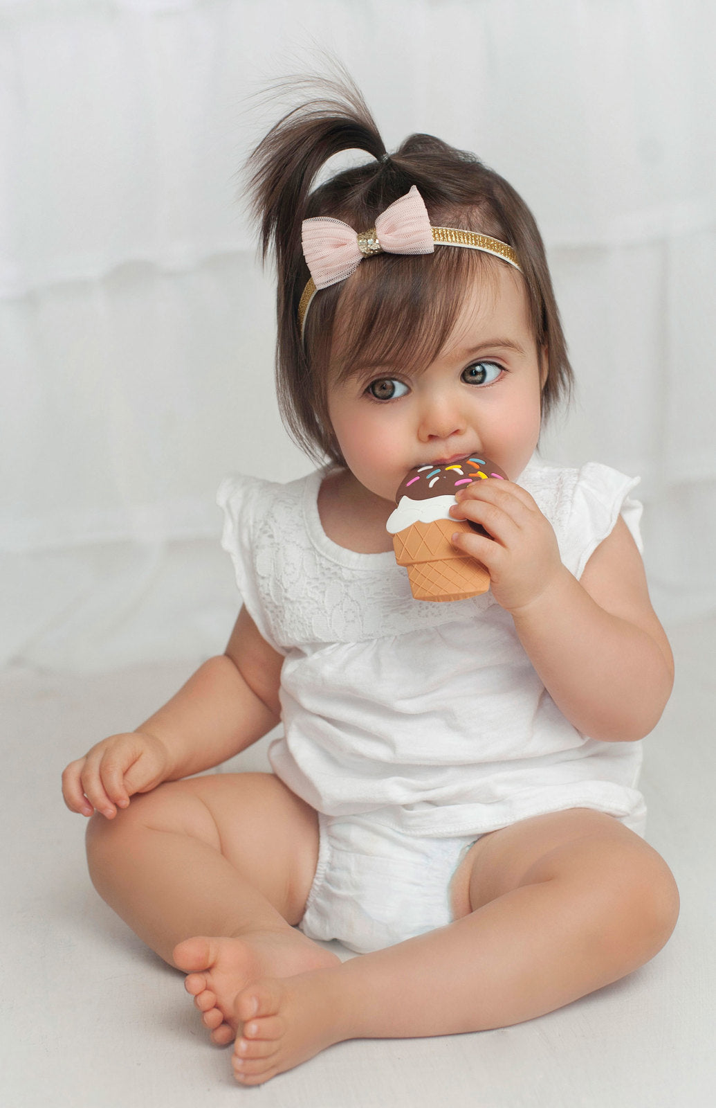 Child in a white outfit with a pink bow eating an ice cream cone on a light background