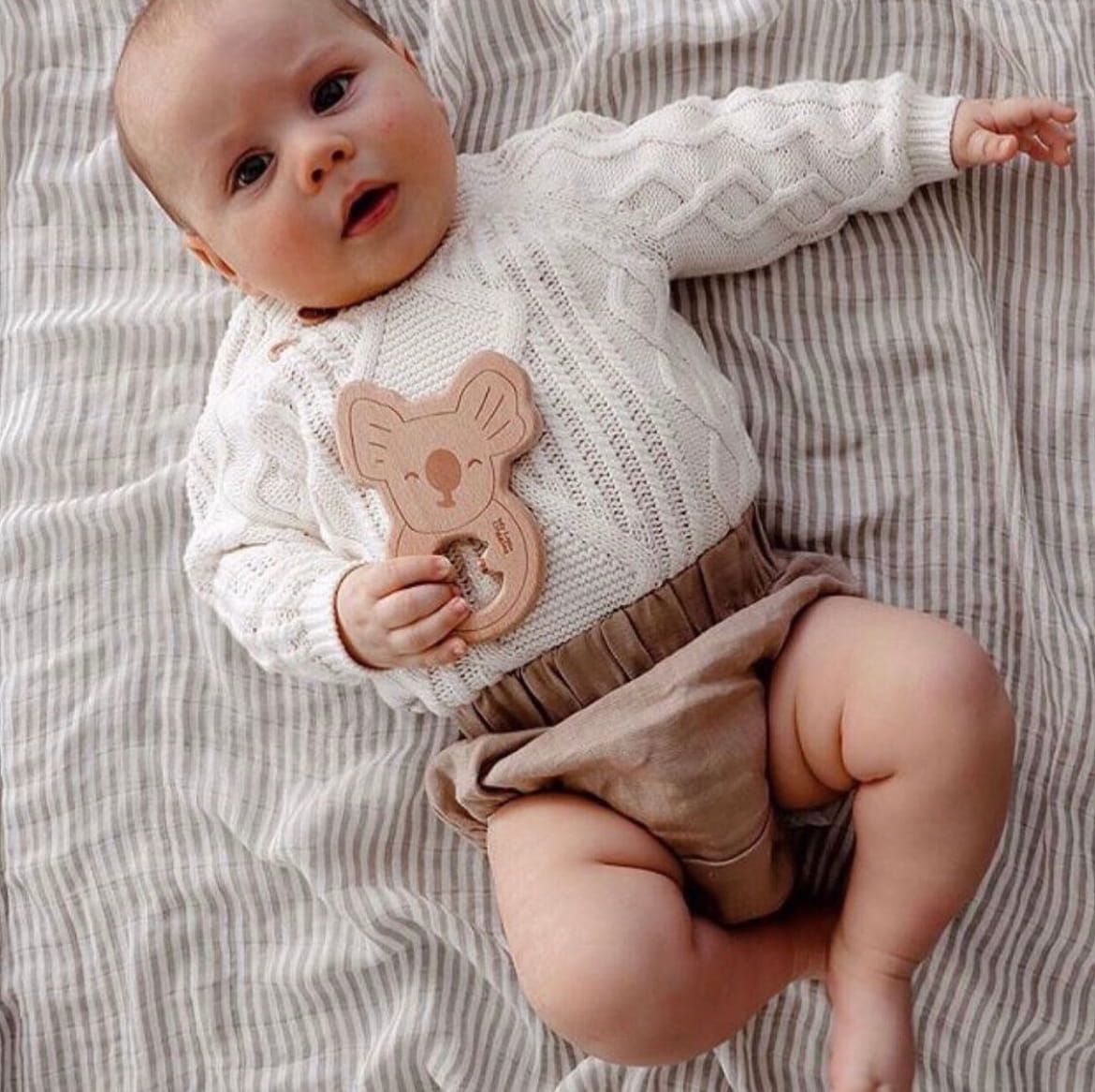 Baby wearing a white cable knit sweater and brown shorts, holding a wooden toy, lying on a striped blanket.