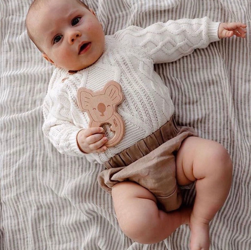 Baby wearing a white cable knit sweater and brown shorts, holding a wooden toy, lying on a striped blanket.