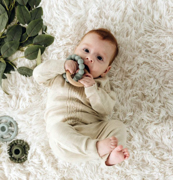 Baby lying on a fluffy white rug with a teething ring, surrounded by green leaves.