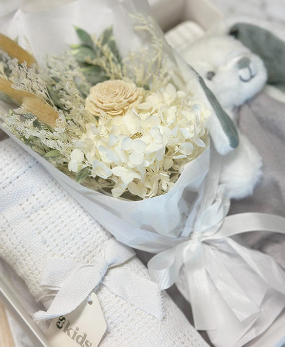 Bouquet of white flowers in a clear vase with a white teddy bear and baby blanket on a light background