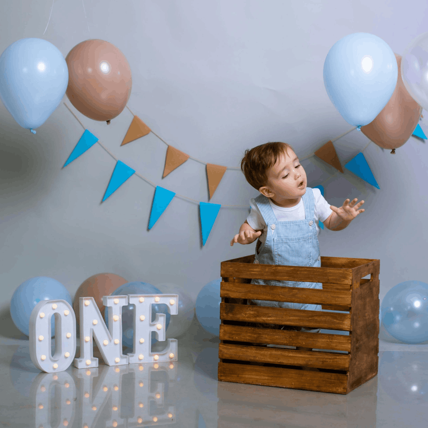 Child in a wooden crate with balloons and 'ONE' letters, celebrating a birthday.