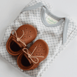 Brown baby shoes with white laces on a patterned grey onesie