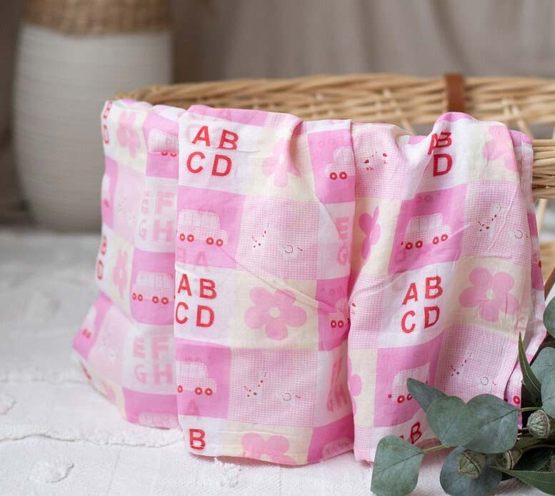 Pink muslin wrap with 'AB CD' text in a basket on a light surface.