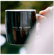 Black mug with humorous text held by a hand against a blurred natural background