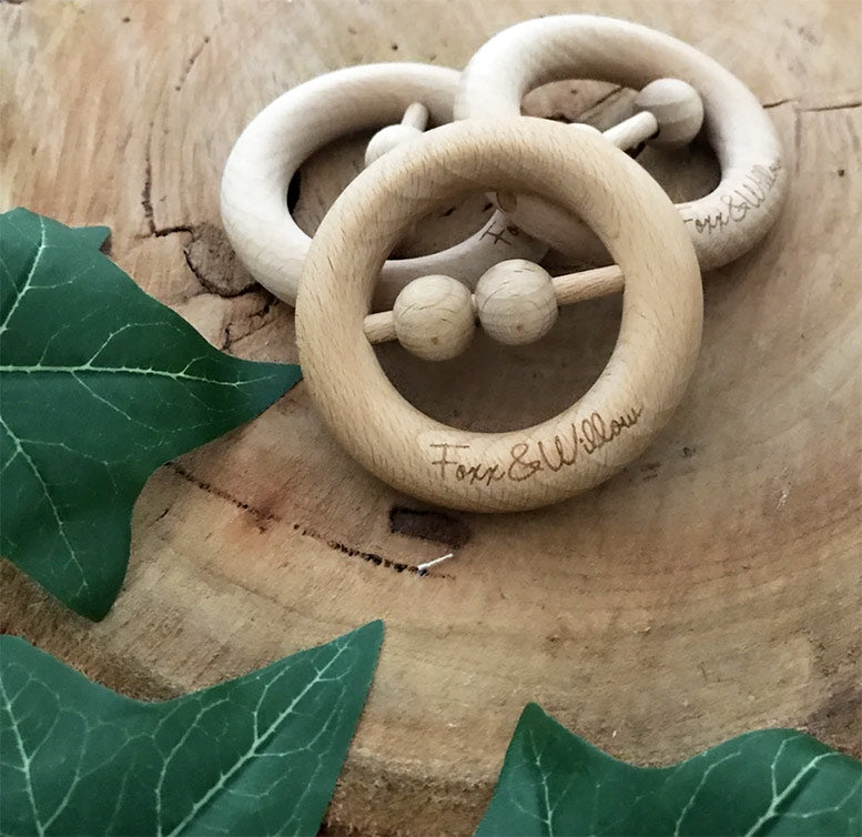 Wooden baby teething ring with 'Fox & Willow' engraving on a wooden surface with leaves.