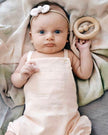 Baby in pink overalls holding a wooden toy, lying on a soft surface.