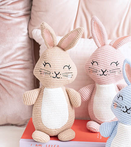 Three crocheted bunny toys on a pink surface with a blurred background