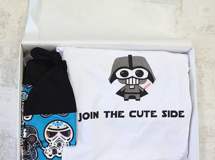 Box containing a black beanie, a white baby onesie with a star wars design, and a baby  hat on a light gray background.