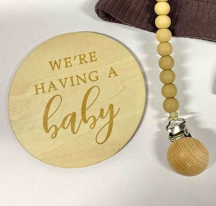 Wooden disc with 'We're having a baby' text and a wooden pacifier clip on a white background.